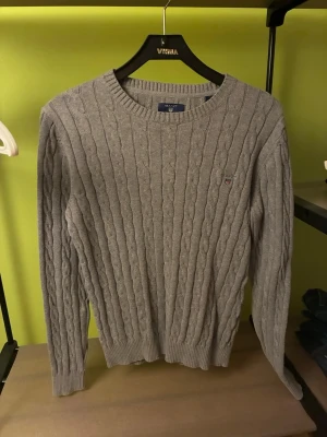 Grå kabelstickad tröja från GANT - Snygg grå kabelstickad tröja från GANT i premium bomull. Tröjan har rund halsringning, ribbade muddar och en liten GANT-logga broderad på bröstet. Perfekt för dig som gillar klassisk och stilren design.
