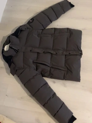 Mörkgrå pufferjacka från Canada Goose - Säljer en mörkgrå pufferjacka från Canada Goose med klassisk patch på ärmen och hög krage. Jackan har långa ärmar, stora fickor och knappar framtill. Perfekt för kalla dagar och har en riktigt schysst oversized look.Fick som present har inte kommit till någon användning. Hör av dig vid frågor elller funderingar.