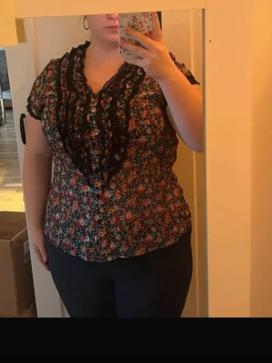 Blommig blus med spetsdetaljer - Söt blommig blus med svarta spetsdetaljer längs urringningen och framtill. Blusen har korta ärmar med liten volangkant och ett färgglatt blommönster i rosa, rött och grönt på svart botten. 
