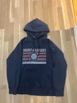 Mörkblå Saint Laurent hoodie - Snygg mörkblå hoodie från Saint Laurent med tryck framtill i vitt och rött samt logga. Klassisk huva med dragsko och stor magficka. Tillverkad i mjukt bomullsmaterial som känns skönt mot huden. Perfekt för en avslappnad och trendig look. Tröjan har aldrig varit använd utan bara provad. Pris kan diskuteras våga ge bud.