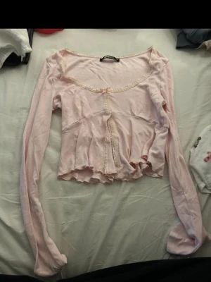 Ljusrosa croppad topp från gina tricot - Söt ljusrosa croppad topp från Gina tricot med långa ärmar, spetsdetaljer längs ringningen och framtill samt en liten rosett vid urringningen. Toppen har volangkant nedtill och är tillverkad i ett mjukt material som känns bekvämt mot huden.