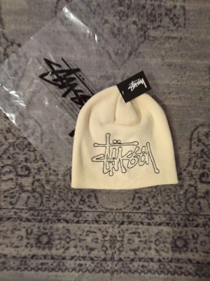 Vit Stüssy mössa med broderad logga - Snygg vit mössa från Stüssy med deras ikoniska broderade logga framtill. Mössan är ribbstickad och har en klassisk passform som passar perfekt till streetwear-stilen. Materialet är mjukt och bekvämt, perfekt för kalla dagar.