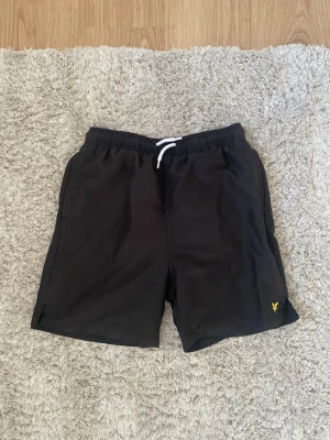 Svarta shorts från Lyle & Scott - Svarta shorts från Lyle & Scott med elastisk midja och vit snörning. Klassisk gul logga broderad på vänster ben. Perfekta för sommaren och har en enkel, stilren look.