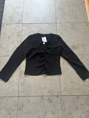 Svart långärmad blus från Bikbok  - Stilren svart blus från Bikbok med lång ärm och snygg rynkning framtill. Blusen har en rund halsringning och är tillverkad i ett mjukt, lätt material som ger en skön känsla. Perfekt att matcha med jeans eller kjol för en trendig look.