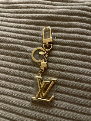 Vuitton guldfärgad nyckelring - Snygg guldfärgad nyckelring från  Vuitton med den ikoniska loggan i metall. Nyckelringen har både ring och karbinhake, samt en liten dekorerad bricka med -gravyr. Perfekt att hänga på väskan eller nycklarna för en lyxig touch.