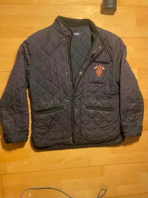 Svart quiltad jacka från Morris - Snygg svart quiltad jacka från Morris med broderad logga på bröstet och två framfickor med dragkedja. Jackan har ståkrage i manchester och stängs med både dragkedja och knappar. Perfekt för dig som gillar klassisk stil.