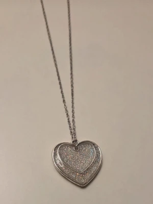 Halsband med dubbla hjärtan i silver glitter rostfritt stål  - Snyggt halsband med två hjärtformade hängen i silverfärg. Båda hjärtana har glittrande yta som ger ett extra lyft till din outfit. Kedjan är tunn och elegant, perfekt för dig som gillar stilrena accessoarer med lite extra bling. Hänge ca 2.5 cm, kedja 45 cm i rostfritt stål. Helt nytt/egentillverkad.