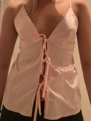 Ljusrosa satintopp - Säljer en ljusrosa linnetopp med smala axelband och djup v-ringning. Toppen har flera snörningar framtill som ger en trendig och feminin look. Perfekt att styla med jeans eller kjol för en cool vibe.
