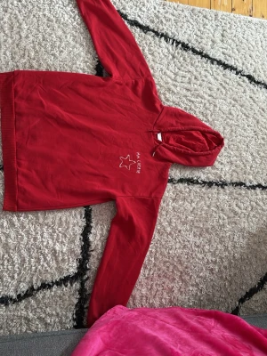 Röd hoodie med tryck Ma Chérie - Snygg röd hoodie med huva och vitt tryck på bröstet där det står 'Ma Chérie' och en stjärna. Den är från Gina Tricot Hoodien har långa ärmar och ribbade muddar. Perfekt för en avslappnad och trendig look. Materialet känns mjukt och bekvämt, troligen bomullsblandning. OBS! Bild 3 visar några fel, det första felet är att den metall saken på snöret är borta på ena och där hoodie kragen slutar är det trasigt men kan sys!