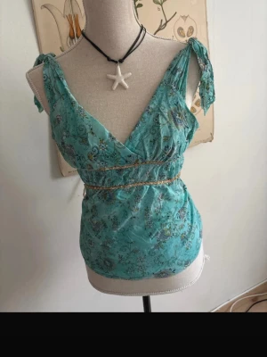 Turkos blommig omlottopp med knyt - Supersöt turkos omlottopp med blommigt mönster och knyt på axlarna. Toppen har v-ringning och smock i midjan för en snygg passform. Perfekt för dig som gillar boho-stil och vill ha något luftigt och färgglatt.