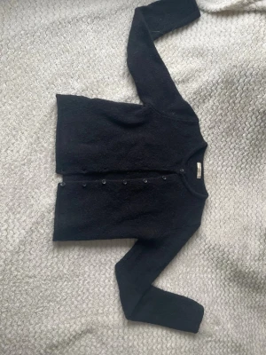 Svart kofta med rund hals - Mysig svart kofta med rund halsringning och knappar framtill. Koftan har lång ärm och är tillverkad i ett mjukt, stickat material som känns skönt mot huden. Enkel och stilren design som passar till det mesta.