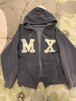 Blå hoodie med dragkedja och tryck - Blå hoodie med dragkedja och stora bokstäver 'M' och 'X' i vitt på framsidan. Utrustad med huva och dragsnören samt två rymliga magfickor. Tillverkad i mjukt bomullsmaterial, perfekt för en avslappnad streetwear-look.