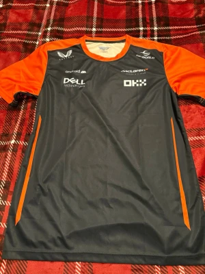 McLaren Norris #4 sporttröja - Snygg sporttröja från McLaren med Norris och nummer 4 på ryggen. Tröjan är svart med orange axlar och detaljer, samt flera sponsorer tryckta på framsidan. Tillverkad i ett lätt och snabbtorkande material, perfekt för träning eller motorsportfans. (Fake)