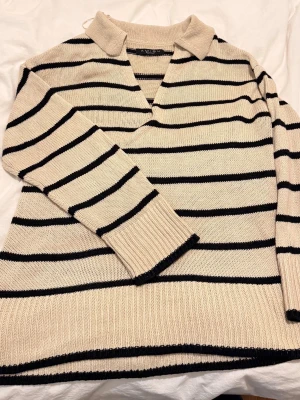 Randig stickad tröja med krage - Säljer en beige stickad tröja från amishu  knitwear med svarta horisontella ränder. Tröjan har en avslappnad passform, krage och v-ringning. Långa ärmar med breda ribbade muddar och ribbad nederkant. Perfekt för en chill och trendig look. Strl S men sitter som M är 164 cm lång