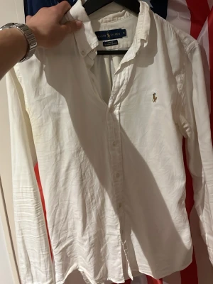Vit skjorta från Ralph Lauren - Klassisk vit skjorta från Ralph Lauren med broderad logga på bröstet. Skjortan har lång ärm, knappar framtill och är tillverkad i mjuk bomull. Perfekt till både jeans och chinos för en clean och stilren look. Slim passform.