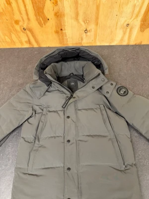 Grå dunjacka från Canada Goose - Snygg grå dunjacka från Canada Goose med avtagbar huva och klassisk patch på ärmen. Jackan har dragkedja och knappar framtill, samt två snedställda fickor med dragkedja. Perfekt för kalla vinterdagar och riktigt varm.