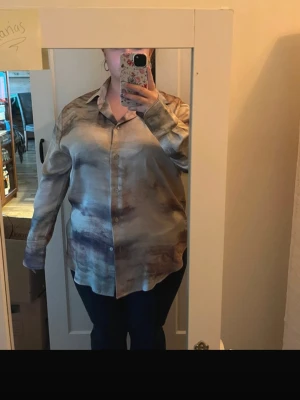 Beige och brun skjorta i satinlook - Säljer en loose fit skjorta i beige och bruna toner med satinliknande material. Skjortan har klassisk krage, långa ärmar och knappar framtill. Perfekt för dig som gillar en avslappnad men ändå stilren vibe. Har ett litet hål i ena armhålan som ej syns 