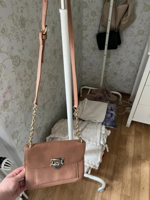 Ljusbrun axelväska med guldkedja - Säljer en snygg ljusbrun axelväska i fejkläder med guldfärgad kedja och spänne med nitar. Väskan har en stilren, rektangulär form och justerbar axelrem. Perfekt för dig som vill ha en trendig och praktisk accessoar till din outfit.
