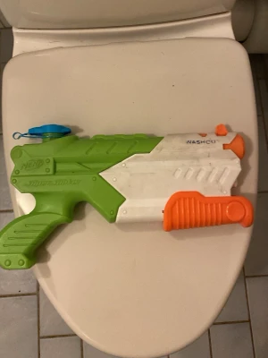 Nerf Super Soaker vattenpistol - Rolig vattenpistol från Nerf Super Soaker-serien i grönt, vitt och orange med blått lock. Pistolen har ett greppvänligt handtag och en pumpmekanism framtill. Perfekt för vattenlekar utomhus under varma dagar.
