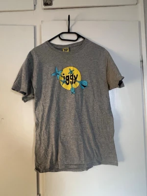 Grå t-shirt från Iggy med tryck - Säljer en grå t-shirt från Iggy med ett stort tryck av blåa pilar och gul cirkel med texten 'iggy' på bröstet. T-shirten har rund hals och är kortärmad. Perfekt för dig som gillar streetwear och coola prints.