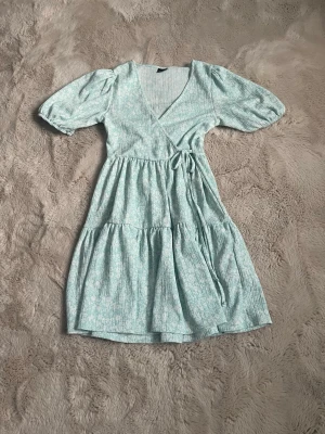 Ljusblå omlottklänning Gina Tricot - Supersöt ljusblå omlottklänning från Gina Tricot med puffärmar och knyt i midjan. Klänningen har ett diskret blommigt mönster, volangdetaljer och v-ringad hals. Perfekt för varma dagar och ger en fräsch sommarkänsla.