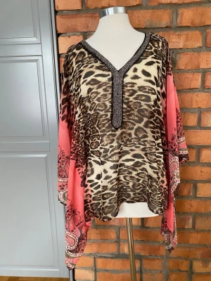 Leopardmönstrad blus med paisley - Säljer en luftig blus med leopardmönster i beige och brunt, samt rosa paisleydetaljer på ärmarna. Blusen har v-ringning med dekorativa pärlor längs kanten och vida, trekvartsärmar. Perfekt för dig som vill sticka ut med ett djurmönster och coola detaljer.