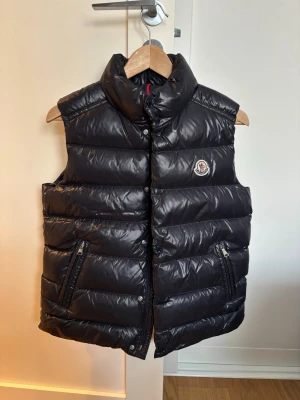 Mörkblå  dunväst från Moncler - Snygg mörkblå dunväst från Moncler med hög krage och två dragkedjefickor framtill. Västen har ett quiltat mönster och klassisk Moncler-logga på bröstet. Tillverkad i glansigt material som ger en exklusiv känsla. Perfekt för lager-på-lager och streetstyle.