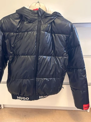 Svart pufferjacka från HUGO - Snygg svart pufferjacka från HUGO med glansig finish och huva. Jackan har dragkedja framtill och elastisk nederkant med HUGO-logga i vitt. Perfekt för kalla vinterdagar och ger en trendig look. Materialet är syntetiskt och jackan har en boxig passform.