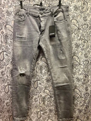 Grå slitna jeans från Dsquared2 - Säljer ett par grå jeans från Dsquared2 med skinny fit och slitna detaljer på benen. Jeansen har fem fickor, dragkedjegylf och coola patchade partier samt råa kanter. Perfekta för en trendig och avslappnad stil.