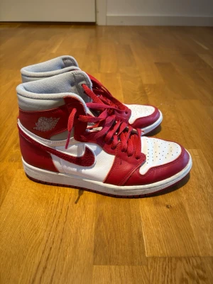 Air Jordan 1 Retro High röd/vit - Säljer ett par Air Jordan 1 Retro High sneakers i rött och vitt med klassisk Nike Swoosh på sidan. Skorna har högt skaft, röda skosnören och detaljer i läder. 