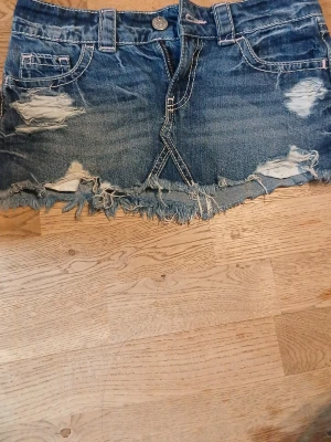 Blå jeanskjol med slitningar gina tricot - Säljer en kort blå jeanskjol med slitna detaljer och fransig nederkant. Kjolen har klassiska fickor fram och bak, bälteshällor och synliga sömmar. Perfekt för dig som gillar en avslappnad och trendig stil.