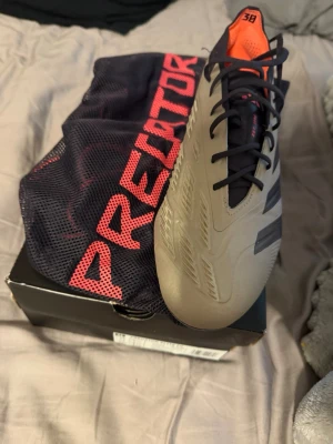 Adidas Predator fotbollsskor beige/svart - Säljer ett par Adidas Predator fotbollsskor i beige med svarta detaljer och orange insida. Skorna har snörning, låg profil och är tillverkade i syntetmaterial för bra bollkontroll. Medföljer originalkartong och svart meshpåse med röd text.