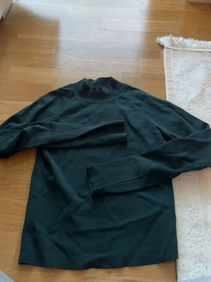 Mörkgrön långärmad tröja - En enkel och stilren mörkgrön långärmad tröja med högre hals. Tröjan har rak passform och är tillverkad i ett mjukt material som passar perfekt till lager på lager. Inga synliga loggor eller mönster, bara clean och snygg.