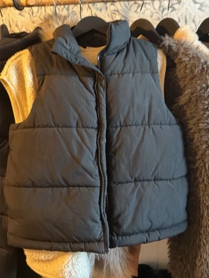 Svart puffig dunväst - Säljer en svart, puffig dunväst med hög krage och dragkedja framtill. Västen har en boxig passform och breda horisontella sömmar som ger en cool, trendig look. Perfekt att slänga över en hoodie eller tröja för extra värme.