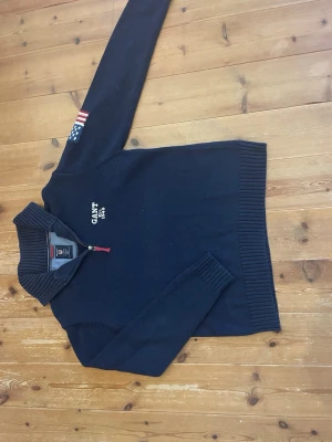 Mörkblå stickad tröja från Gant - Snygg mörkblå stickad tröja från Gant med halv dragkedja och hög ribbad krage. Broderad Gant-logga och 'Est. 1949' på bröstet samt amerikansk flagga på ärmen. Perfekt för dig som gillar klassisk och sportig stil.