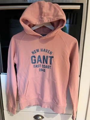 Rosa hoodie från GANT med tryck - Säljer en rosa hoodie från GANT med blått tryck på bröstet där det står 'NEW HAVEN GANT EAST COAST 1949'. Hoodien har huva, känguruficka och ribbade muddar. Perfekt för en chill och sportig look.