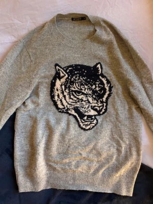 Grå stickad tröja Tiger of Sweden - Stickad grå tröja från Tiger of Sweden med ett stort svartvitt tigerhuvud på bröstet. Tröjan har rund halsringning och ribbade muddar vid ärmslut och nederkant. Perfekt för dig som vill ha en cool statement-piece i garderoben.