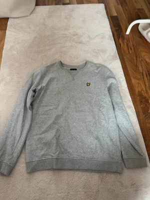 Grå sweatshirt från Lyle & Scott - Klassisk grå sweatshirt från Lyle & Scott med rund halsringning och den ikoniska gula örnloggan broderad på bröstet. Tröjan har ribbade muddar vid ärmslut och nederkant. Perfekt basplagg för en avslappnad stil.