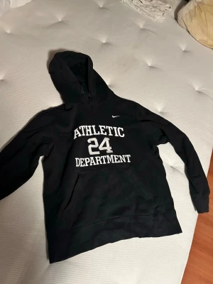 Svart Nike hoodie Athletic Dept 24 - Svart hoodie från Nike med vit text 'ATHLETIC DEPARTMENT 24' på bröstet och klassisk Nike-logga. Hoodien har huva och en stor magficka framtill. Perfekt för en sportig och avslappnad stil.