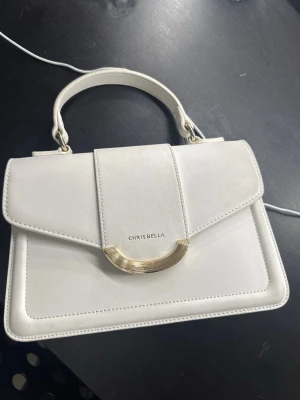 Vit handväska  - Stilren vit handväska med elegant gulddetalj på framsidan. Väskan har ett topphandtag och en strukturerad, rektangulär form. Tillverkad i slätt konstläder och har en klassisk, minimalistisk look.