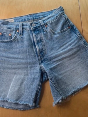 Levi's ljusblå jeansshorts - Klassiska ljusblå jeansshorts från Levi's med rå kant och femficksmodell. Shortsen har hög midja, knappgylf och tydliga Levi's-detaljer som bakfickor med söm och läderpatch bak. Perfekta för sommaren och tillverkade i slitstark bomull.