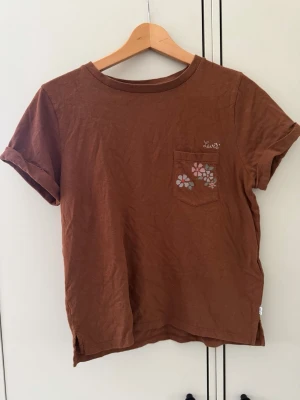 Brun Levi's t-shirt med blomdetalj - Snygg brun t-shirt från Levi's med korta ärmar och en liten bröstficka med broderade blommor. Tillverkad i mjuk bomull och har en avslappnad passform. Perfekt för en chill och stilren look.