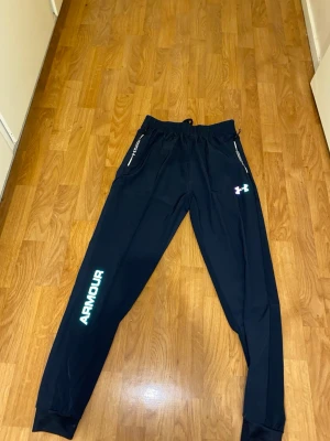 Svarta träningsbyxor Under Armour - Snygga svarta träningsbyxor från Under Armour med resår i midjan och dragsko. Byxorna har vita och gröna logotyper på benet och vid fickan, samt dragkedjeförsedda fickor. Använda 0 ggr