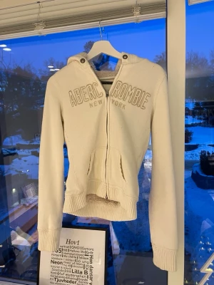 Vit hoodie från Abercrombie - Snygg vit hoodie från Abercrombie & Fitch med dragkedja och broderad logga på bröstet. Hoodien har en mysig fuskpälsfodrad huva och  fickor framtill. ger en skön passform. Perfekt för kalla dagar och är i storlek M men passar säkert 12-15!🤍