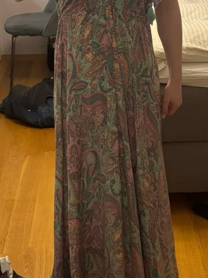 Mönstrad långklänning med snörning - Långklänning i bohemisk stil med paisley- och blommönster i grönt, rosa och beige. Klänningen har snörning i ryggen och omlott-detalj framtill. Ärmlös design och luftig passform, perfekt för sommarens varma dagar.