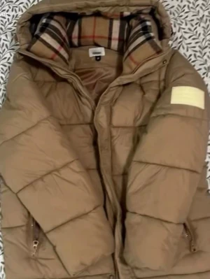 Beige dunjacka från Burberry  - Snygg beige dunjacka från Burberry med klassiskt rutigt foder i huvan. Jackan har dragkedja, två fickor med dragkedja och en patch på ärmen. Perfekt för kalla dagar och ger en stilren look med exklusiv känsla.
