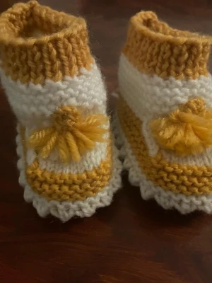 Stickade babytofflor i gult och vitt - Supersöta handstickade babytofflor i vitt och senapsgult garn. Tofflorna har dekorativa tofsar framtill och ribbstickad kant upptill för extra mysfaktor. Perfekta för att hålla små fötter varma och stiliga.