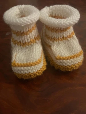Stickade tofflor i vitt och gult - Säljer ett par ny  handstickade tofflor i vitt och gult. De har en mjuk och fluffig känsla med ribbad kant och dekorativa ränder. Perfekta för att hålla fötterna varma hemma. Materialet är garn, troligtvis akryl eller ullblandning.