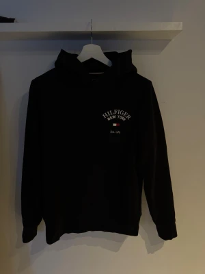Svart hoodie från Tommy Hilfiger - Snygg svart hoodie från Tommy Hilfiger med broderad logga och texten 'Hilfiger New York' på bröstet. Klassisk huva och ribbade muddar vid ärmar och midja. Tillverkad i mjukt bomullsmaterial för en skön känsla.