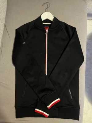 Svart zip hoodie Superdry - Snygg svart hoodie från Superdry med dragkedja framtill och två fickor med dragkedja. Detaljer i rött och vitt längs dragkedjan och på muddarna. Tillverkad i mjukt material, perfekt för en avslappnad stil. Klassisk passform utan huva.